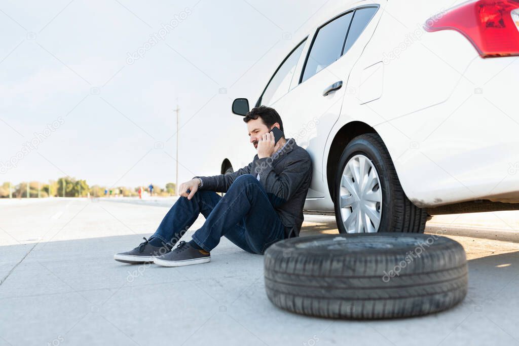 Hispanic man calling a tow truck service because of a flat tire. Man sitting on the road next to his broken-down car 