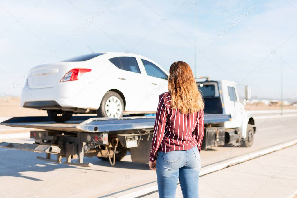 Rear view of a blond young woman watching her car being towed on a tow truck on the side of the highway 