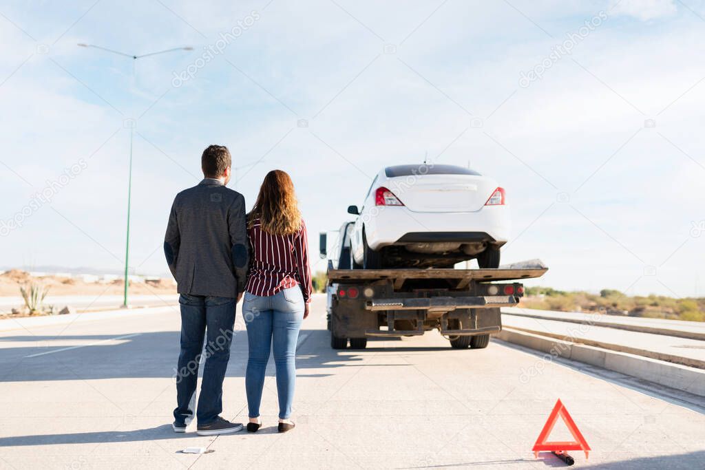 A man in his 30s and a young woman seen from behind and looking sad. Couple watching their car being towed in a tow truck