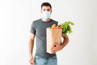 Yüzü maskeli İspanyol genç adam Coronavirus salgını sırasında sebze ve meyve dolu bir market çantası taşıyor.