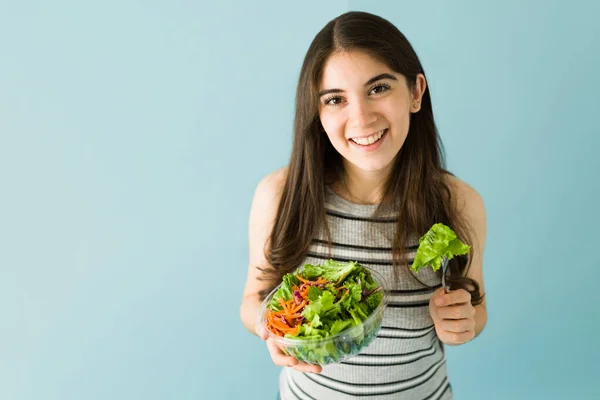 一个20多岁的迷人的微笑的女人用叉子吃绿色沙拉的画像 工作室在蓝色背景下拍摄 — 图库照片
