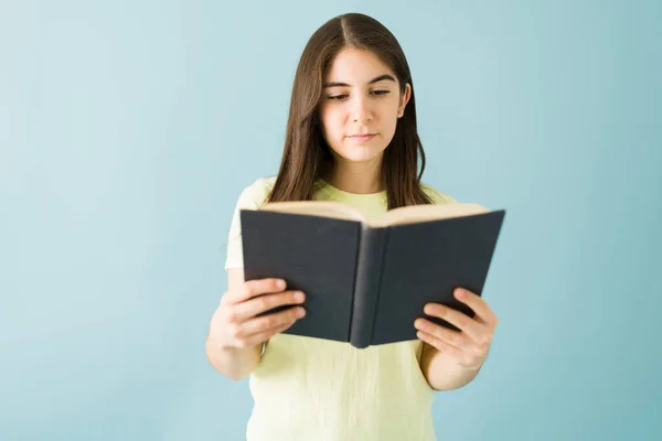 年轻的女学生拿着一本书 带着兴趣的目光在蓝色背景面前看书 — 图库照片