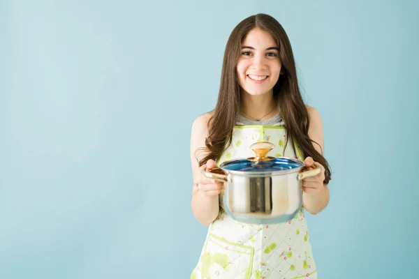 一个笑容满面的年轻女子穿着围裙 在蓝色背景面前端着一个汤壶的画像 — 图库照片