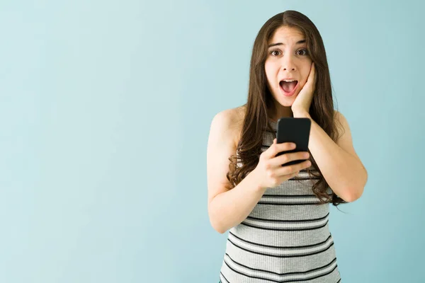 在蓝色背景面前 一个拿着智能手机的白人年轻女人带着惊讶和震惊的表情 — 图库照片