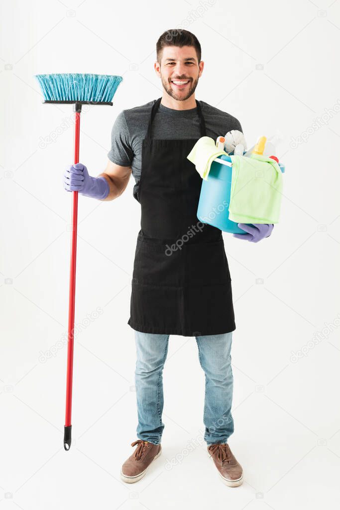 Good-looking young man is smiling and wearing an apron and gloves. Guy holding a broom and carrying a bucket with cleaning products 