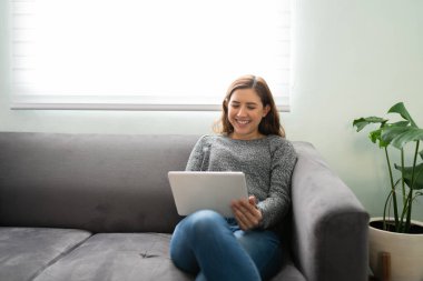 20 'li yaşlarda mutlu bir kadın kanepede oturuyor ve oynuyor, tabletinde online videolar izliyor.