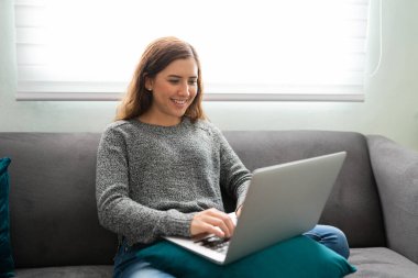 Oturma odasında otururken dizüstü bilgisayarında yazan gülümseyen beyaz bir kadın. 