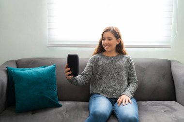 20 'li yaşlarda güzel bir kadın oturma odasında akıllı hoparlörünü tutarken mutlu görünüyor.