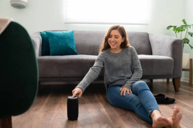 Gülümseyen genç bir kadın oturma odasının zemininde akıllı hoparlörünü açıyor.