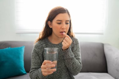 20 'li yaşlarda güzel bir kadın oturma odasında bir bardak suyla hap içiyor.