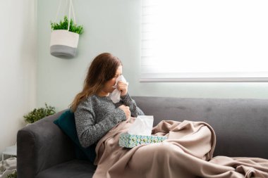 Yirmili yaşlarda bir sarışının kanepeye uzanmış battaniyeli görüntüsü. Hasta kadın soğuk algınlığı ya da grip olduğu için burnunu mendille siliyor. 