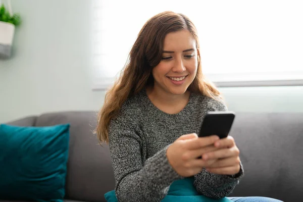 Beautiful young woman sending a text on her smartphone and smiling while sitting on the couch