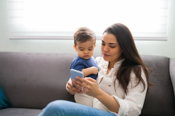 美しいです若いです母とともに彼女の小さな息子座っています上のソファ使用スマートフォン — ストック写真