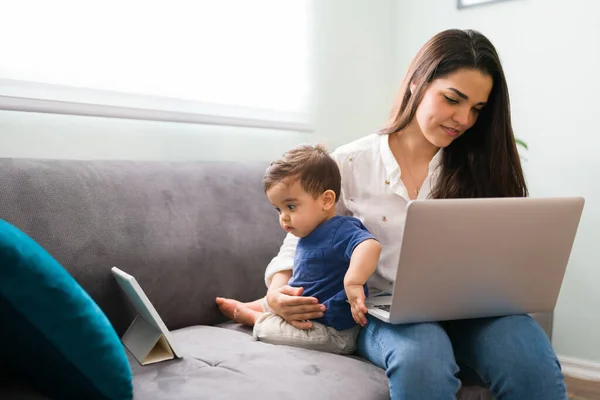 Pretty young single mom working on a laptop while her baby boy looking at digital tablet