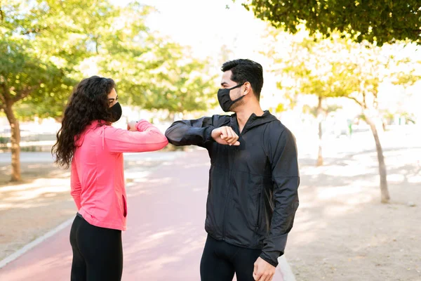 Hispanic young woman and latin young man with face masks and sportswear are greeting each other with an elbow bump in the park because of the coronavirus pandemic