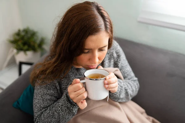 彼女はインフルエンザや風邪で病気ですので 彼女の20代の彼女のソファに座って お茶の熱いカップを飲むの病気の女性のトップビュー — ストック写真
