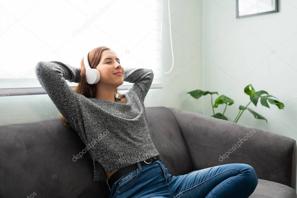 Caucasian young woman looking happy and peaceful while sitting in her living room couch and listening to music with headphones