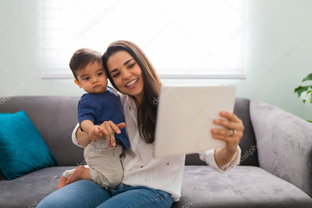 Cute toddler with her gorgeous mother using a digital tablet during a video call with family while sitting on sofa at home