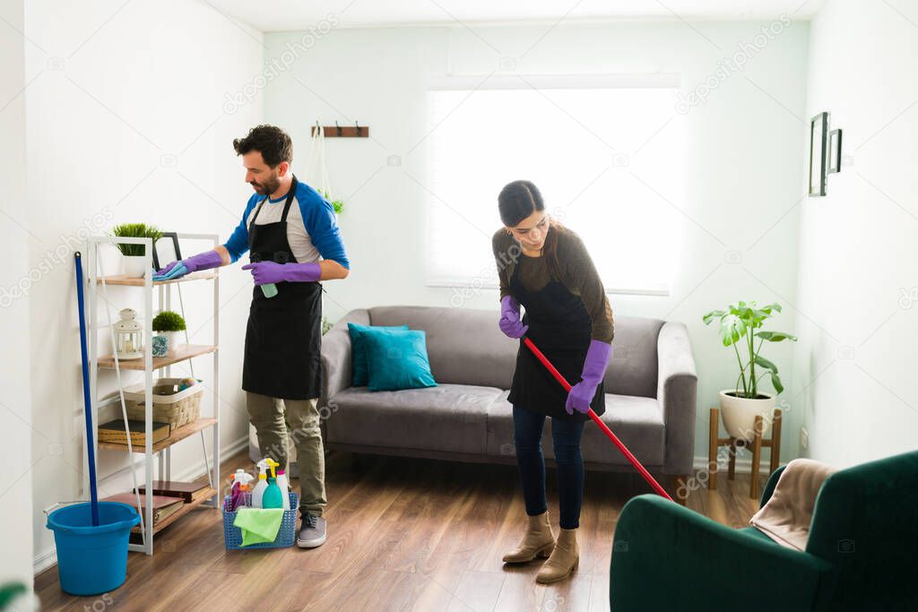 Full length view of a young couple cleaning their home. Man dusting and wiping surfaces while woman sweeps the floor