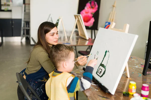 西班牙裔女教师指示一个高加索裔小男孩用画笔在空白的画布上画画 准备上美术课 — 图库照片