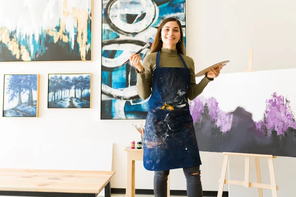 快乐的年轻女画家站在她的艺术工作室里 手里拿着画笔和调色板 女人在新的未完成的画布旁边显得很自豪 — 图库照片