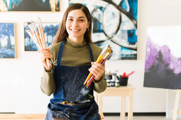 用围裙向年轻的艺术家微笑 拿着很多画笔 漂亮的女画家坐在艺术工作室的长椅上 — 图库照片