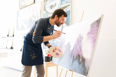 Yakışıklı yetişkin erkek ressam, elinde palet tutarken fırçayla büyük bir tuval üzerinde yeni bir sanat çalışması yapıyor. 