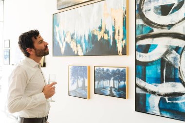 30 'larında yakışıklı bir adam sanat galerisinde sergilenen tablolara hayran. Bir bardak şaraplı erkek ressam diğer sanatçıların resimlerine bakıyor.  