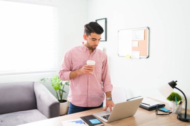 Yönetici olarak çalışan yakışıklı genç adam elinde kahve tutarken bilgisayarında e-postalarını kontrol ediyor. 