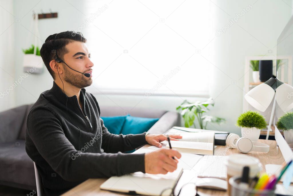 Latin man in his 20s sitting at his desk and wearing a headset. Attractive guy working as a translator with a notebook and books in his workspace