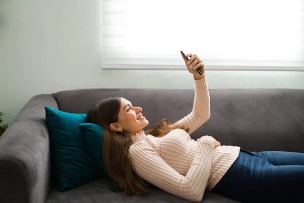 20多岁的女人笑着躺在沙发上 一边在智能手机上与她的长距离男友聊天 一边放松自己 — 图库照片