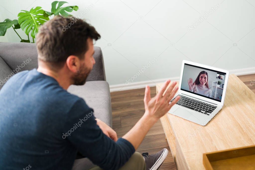 Happy latin man sitting on the couch with his laptop and waving hello to his smiling long-distance girlfriend during an online date 