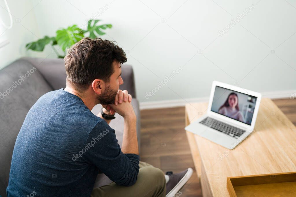 Rear view of a hispanic boyfriend sitting on the living room talking with his pretty girlfriend during a video-call on his laptop