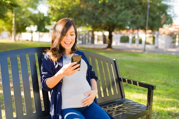 スマホでメールをしながら笑っている妊婦さんの興奮 魅力的な白人女性が休憩して公園のベンチに座って — ストック写真