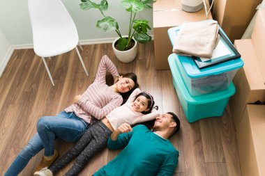 Genç bir aile yukarıda yerde yatarken ve mobilyalarını boşaltırken görülmüş. Mutlu genç kadın, erkek ve küçük kız yeni bir eve taşındıktan sonra mola veriyorlar.