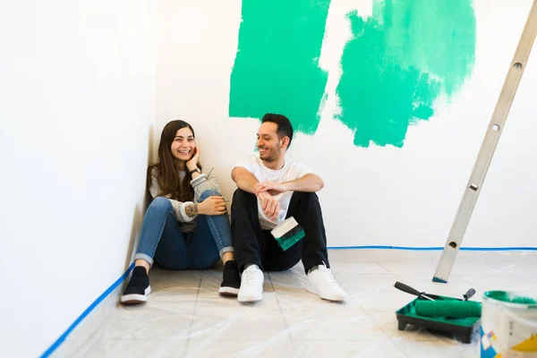 かなりのガールフレンドとボーイフレンドが壁をペイントし 彼らの新しいアパートを改装から休憩を取ります 魅力的なカップル座っている彼らの新しい家の床 — ストック写真