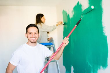 Yakışıklı bir adam kameraya bakıyor ve elinde boya silindiri tutarken gülümsüyor. Latin genç kadın duvarı fırçayla parlak yeşile boyuyor. 