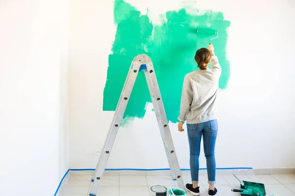 はしごの横に立ってから見て 彼女の新しいアパートや家の壁にローラーで緑の塗料を適用する若い女性 — ストック写真