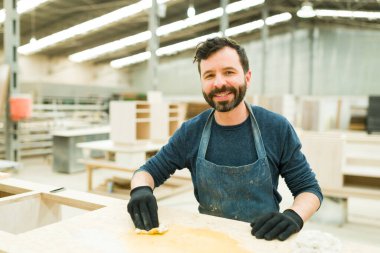 Portrait of a smiling carpenter staining wood with oil stain in his big workshop. Hard-working worker doing his job in a wood shop clipart