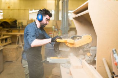 Testereyi kullanırken koruyucu ekipman takan profesyonel bir erkek marangoz. İspanyol adam bir parça odun kesiyor ve talaşla kaplanıyor. 