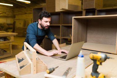 Önlüklü yakışıklı marangoz büyük bir atölyede dizüstü bilgisayarla çalışıyor. Erkek işçi internetten marangozluk ipuçları arıyor