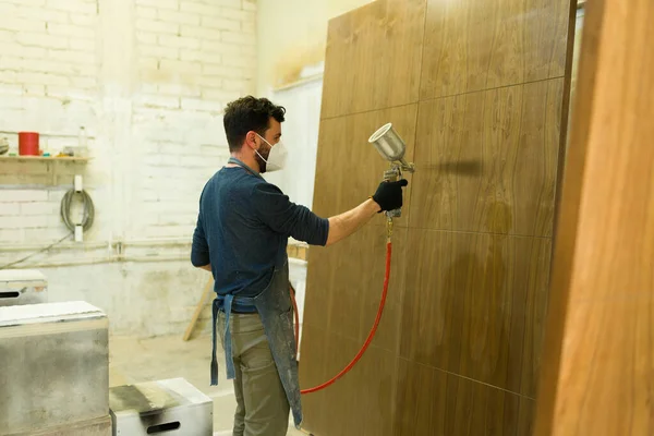 带着手套和面罩的拉丁男工拿着喷雾枪 在一家木材店里画着棕色木门 — 图库照片