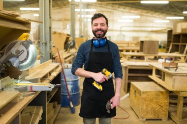 Smiling hispanic man doing carpentry work in a woodshop. Happy carpenter working with a power drill and a hammer clipart