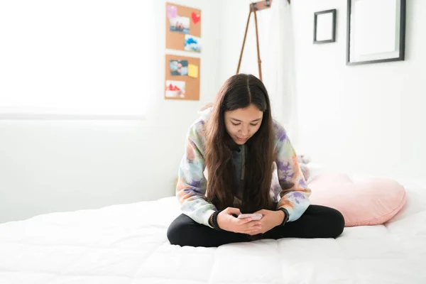 漂亮的少女盘腿坐在床上 低头看她的智能手机 快乐的青少年使用社交媒体和短信在她的手机 — 图库照片