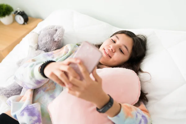 快乐的少女躺在床上 抱着一个蓬松的枕头 漂亮的高加索少女看着社交媒体 用智能手机发短信 — 图库照片