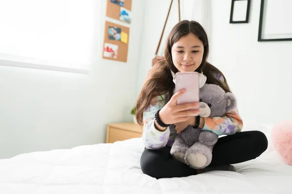 快乐的少女坐在床上抱着一只泰迪熊 漂亮的少女在智能手机里看照片和社交媒体 — 图库照片