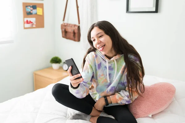 有趣的少女拿着智能手机在卧室里自私自利 白人少女用舌头做鬼脸 — 图库照片