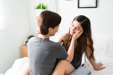 Güzel beyaz bir kadın kız arkadaşının yüzünü okşuyor ve ona dokunuyor. Lezbiyen eşcinsel eşler yatak odalarında yan yana otururlar.