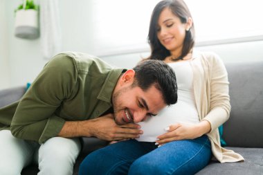 Yakışıklı çekici bir adam karısının karnındaki bebeğin kalp atışlarını dinliyor. Kocası kulağını hamile bir kadının karnına dayamış.