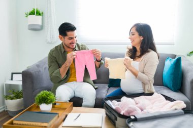 Mutlu yakışıklı adam ve güzel hamile kadın kanepeye otururken bebek kıyafetlerini hastane çantasına koyuyorlar.